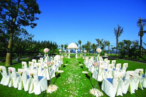 An outdoor wedding aisle with white chairs and tables, a white gazebo, and palm trees.