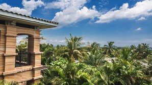 Un bâtiment en bois doté d’un balcon, entouré d’une végétation tropicale luxuriante et de palmiers, sous un ciel bleu dégagé.