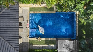 An aerial view of a rectangular swimming pool with a tiled floor, surrounded by a low stone wall and lush greenery.