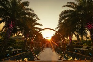 A pathway lined with palm trees and illuminated arches leading to a pool area.