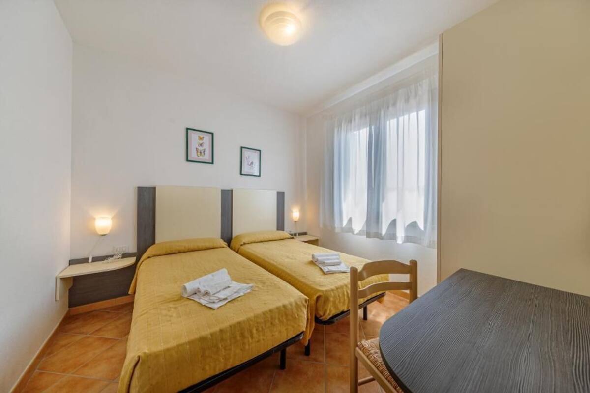 Hotellihuone, jossa on kaksi erillistä sänkyä, puinen tuoli, pieni pöytä ja ikkuna, jossa on verhot.
