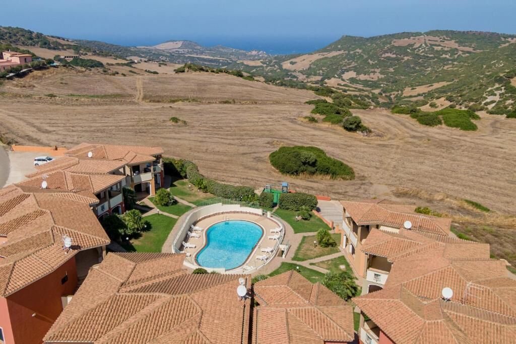 A residential area with terracotta-roofed buildings, a swimming pool, and a view of the sea and hills.