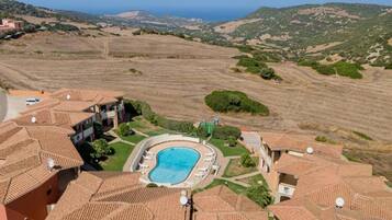 A residential area with terracotta-roofed buildings, a swimming pool, and a view of the sea and hills.