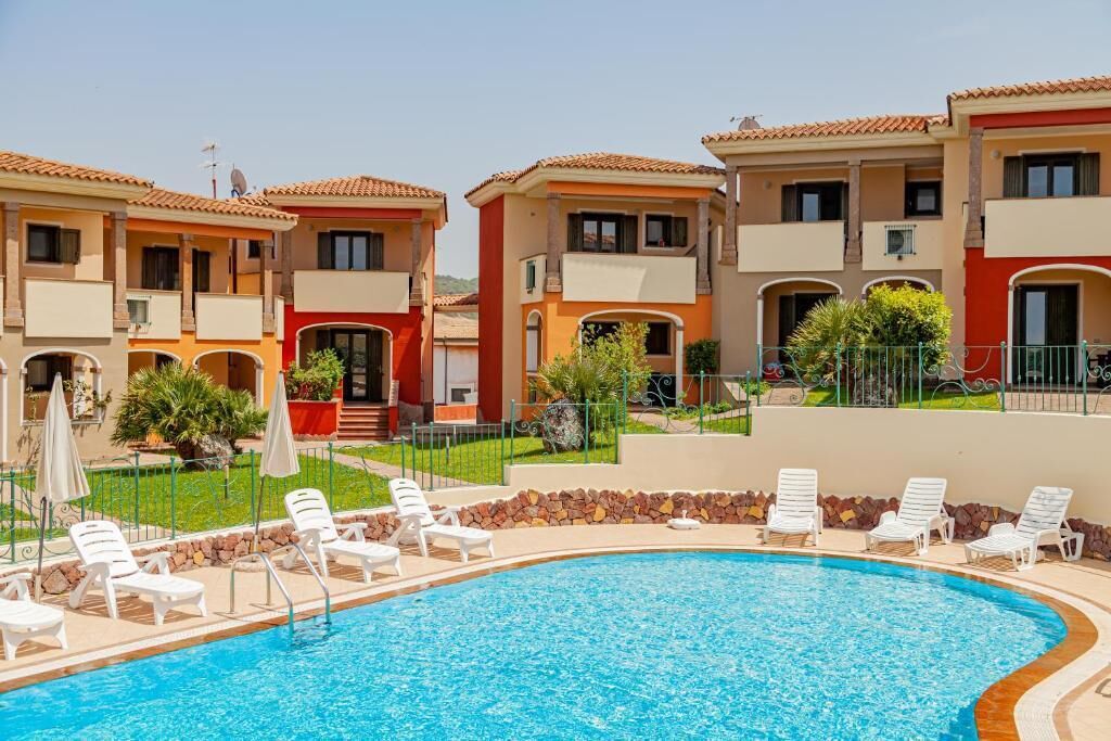 A swimming pool surrounded by white lounge chairs, with a building featuring balconies and a tiled roof in the background.
