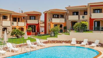 A swimming pool surrounded by white lounge chairs, with a building featuring balconies and a tiled roof in the background.