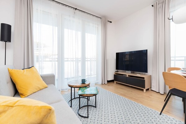 Ein modernes Wohnzimmer mit einer Couch, einem Fernseher, einem kleinen Tisch und einem Fenster mit Vorhängen.