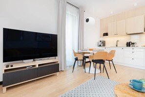 Un salon moderne dotĂ© dâun tĂ©lĂ©viseur Ă Ă©cran plat, dâun coin repas avec des chaises, dâune cuisine avec des placards et dâune grande fenĂȘtre agrĂ©mentĂ©e de rideaux.