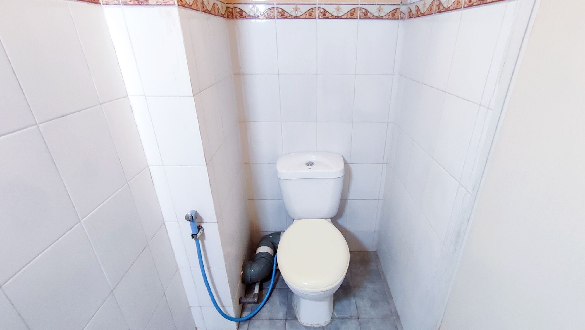 Un baño pequeño con paredes de azulejos blancos, un inodoro y un manguera azul.