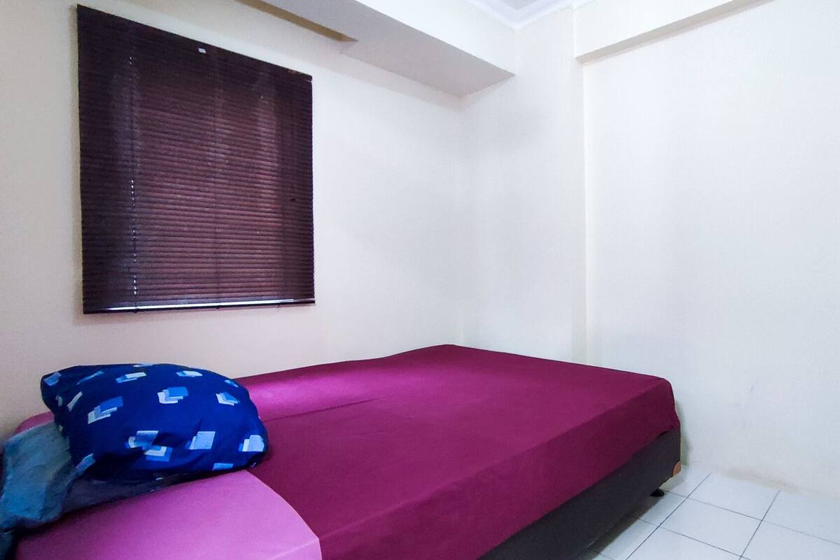 Une petite chambre avec un lit simple, un mur blanc uni, une fenêtre avec des stores et un lit recouvert d’une couverture rouge et agrémenté d’un oreiller bleu.
