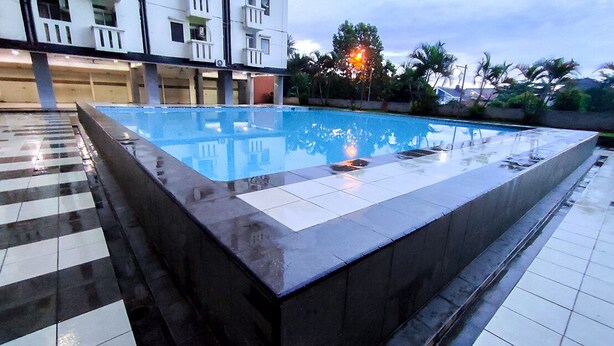 A rectangular pool with a tiled deck surrounding it, a building with balconies in the background, and palm trees. - Lily Room at Apartment Cibubur Village (Depok)