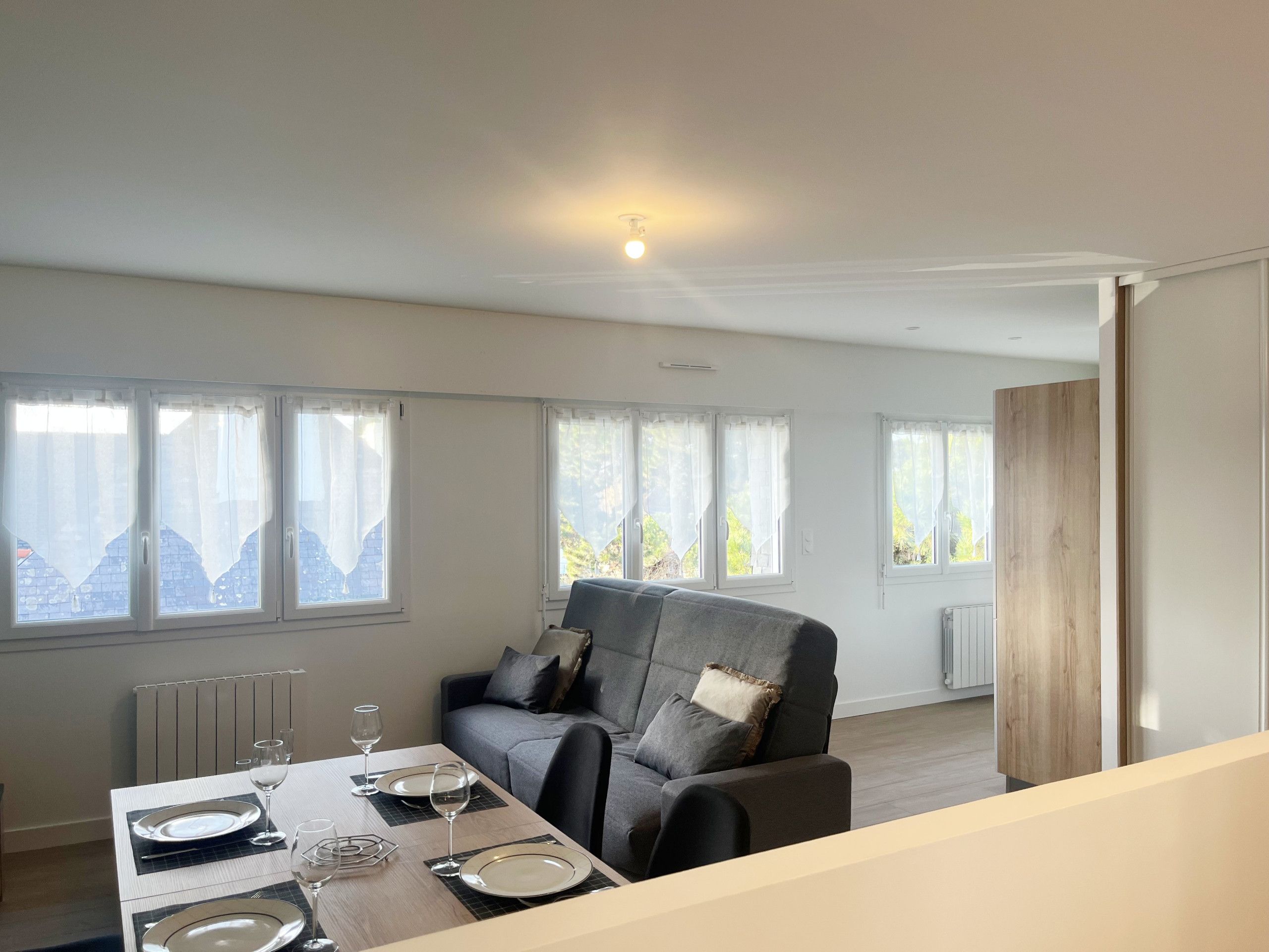 Een eetruimte met een tafel voor vier personen, een grijze bank en uitzicht op het landschap door de ramen.