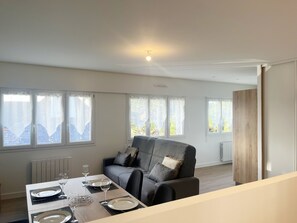 A dining area with a table set for four, a grey sofa, and a view of the outdoors through the windows. - DRUIDES 3 - Holiday rentals - Carnac, Morbihan - T5 (Carnac)