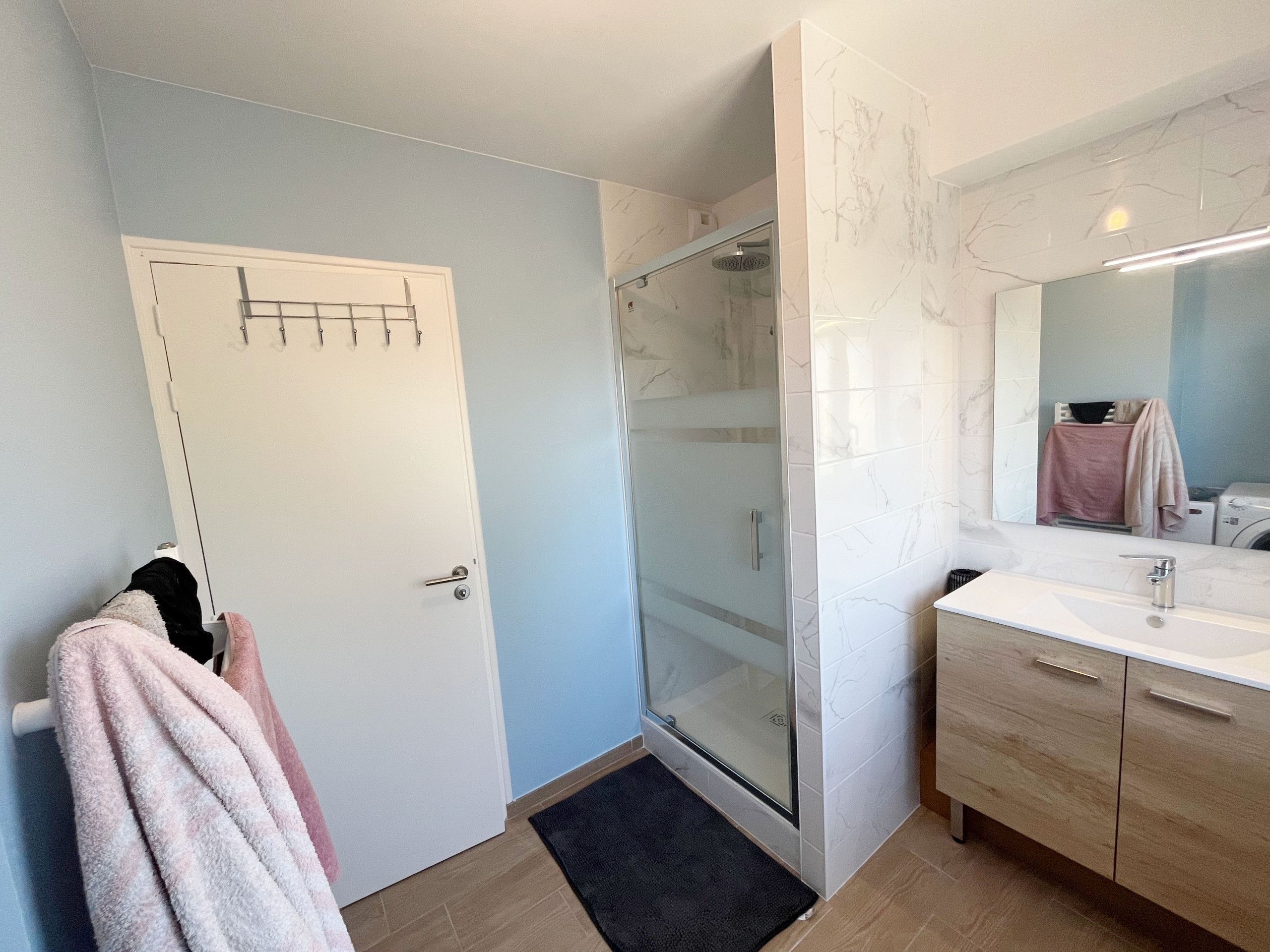 Een badkamer met een glazen douchecabine, een witte deur, een handdoekrek met handdoeken, een wastafel met spiegel en houten kastjes.