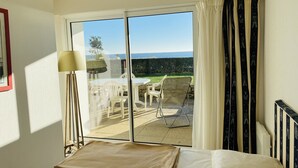 A hotel room with a bed, a table with chairs, a lamp, and a view of the sea through large sliding doors. - HORTENSIAS - Holiday apartment with sea view, facing the beach - T6 (Carnac)