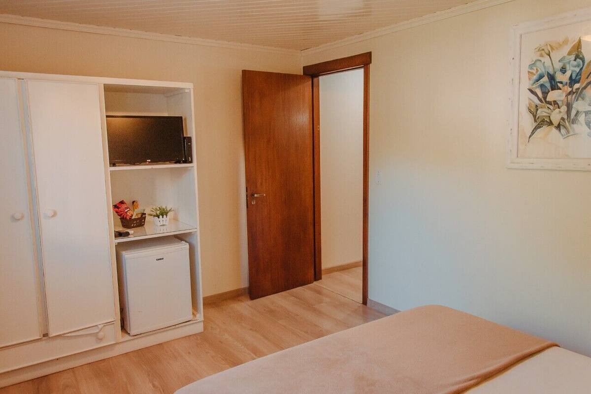 A bedroom with a bed, a wardrobe, a small fridge, and a door leading to another room.