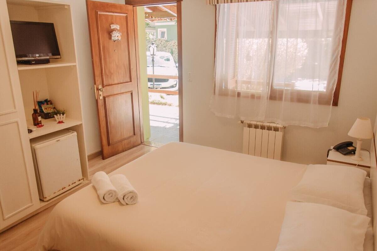 A hotel room with a bed, a TV on a shelf, a radiator, a window with sheer curtains, and a door leading outside.