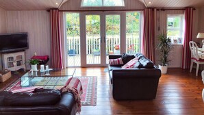 Een ruime woonkamer met een grote televisie, leren banken, een glazen salontafel en uitzicht op een houten terras door glazen deuren.
