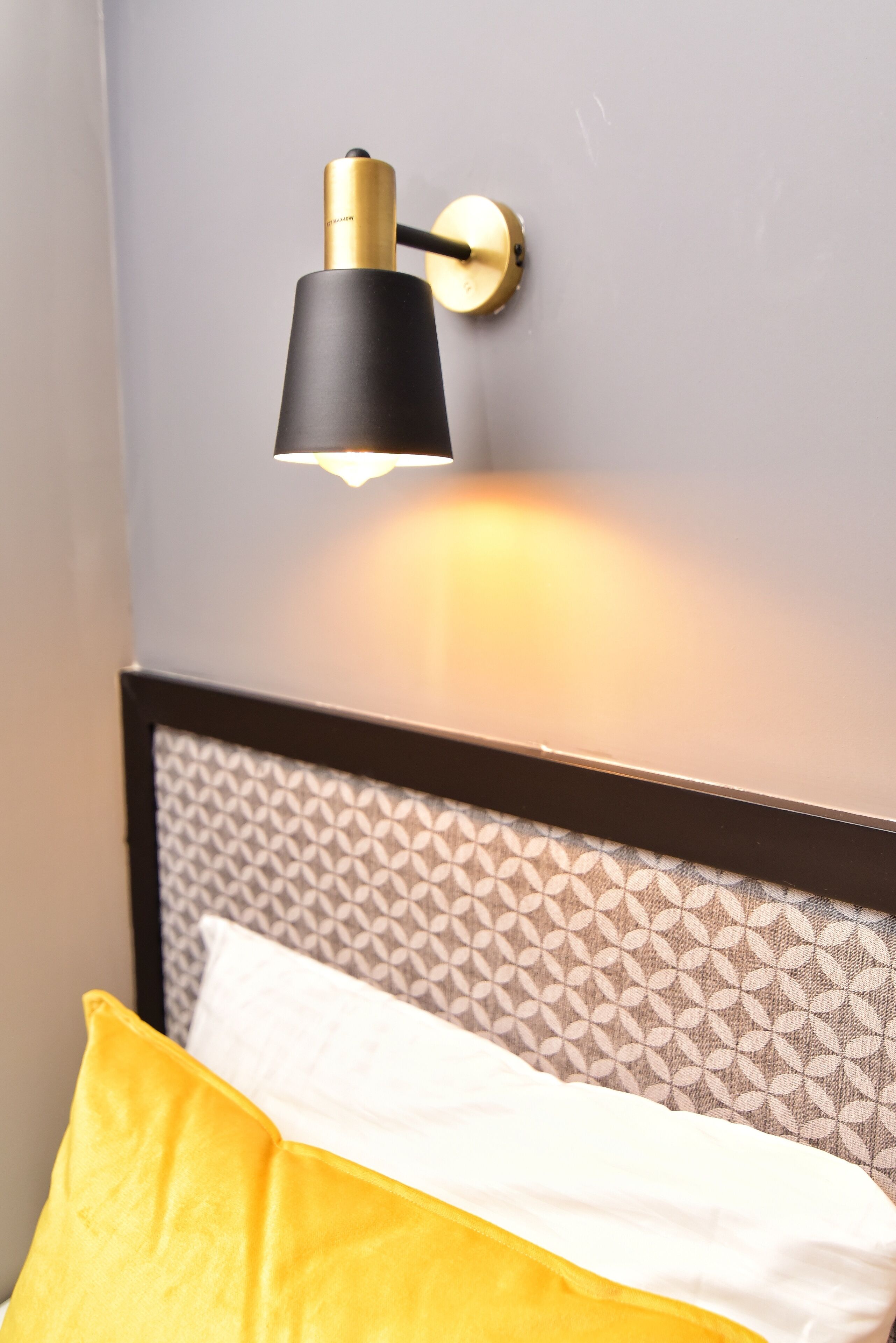 Un lit avec une tête de lit à motifs, une lampe fixée au mur et un coussin jaune.