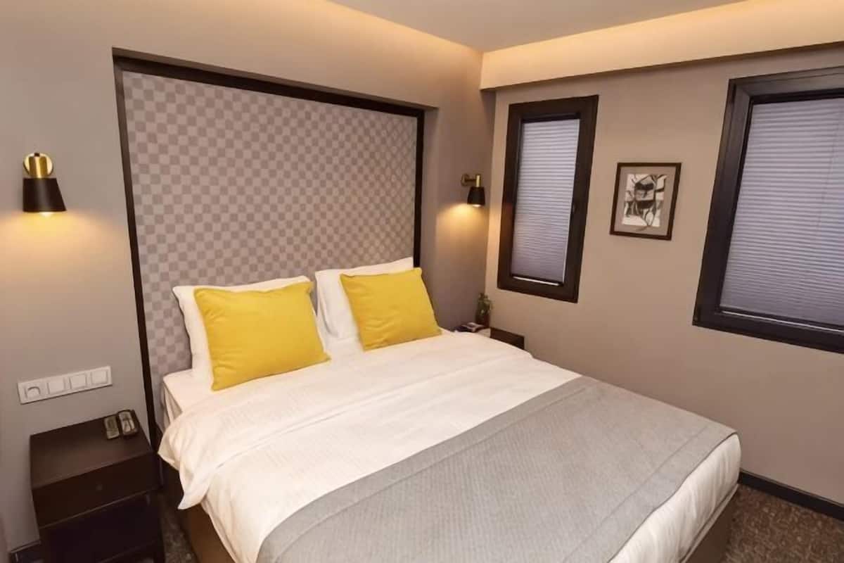 A hotel room with a large bed, two bedside lamps, a nightstand, and a window with blinds.