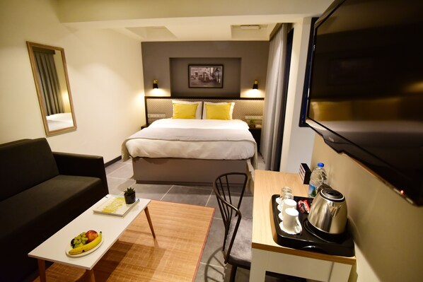 Une chambre d’hôtel comprenant un lit, un bureau avec une bouilloire, une machine à café, un panier de fruits, un canapé et une télévision.