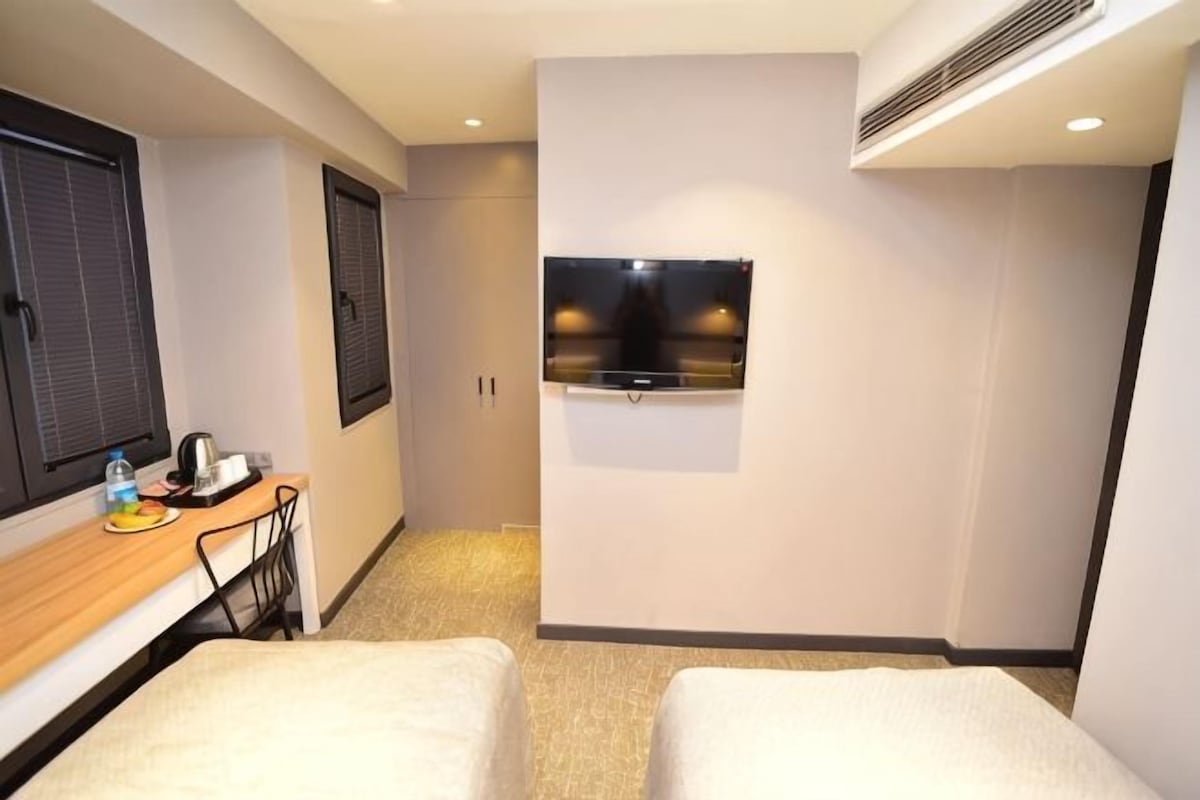 Une chambre d’hôtel avec deux lits, un bureau avec une chaise, une télévision et une fenêtre avec des stores.