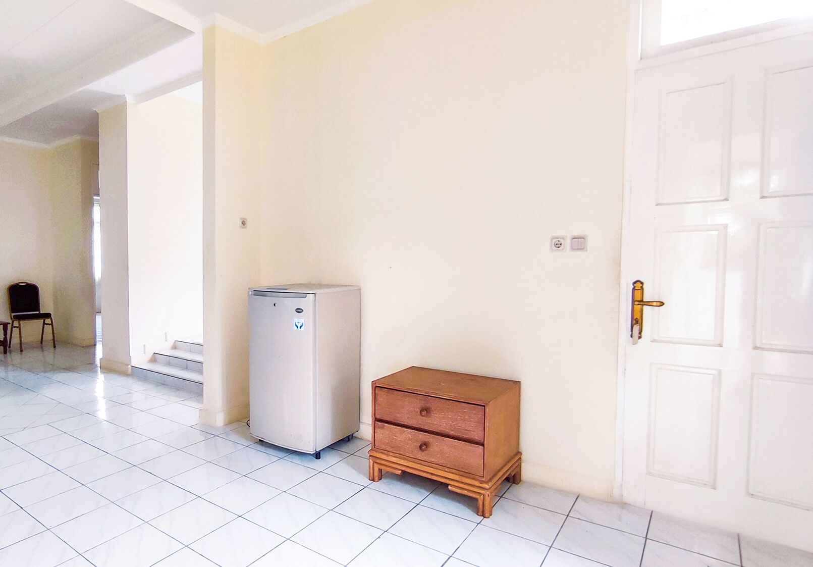 Una stanza con una porta bianca, un piccolo comò di legno e un frigorifero bianco.