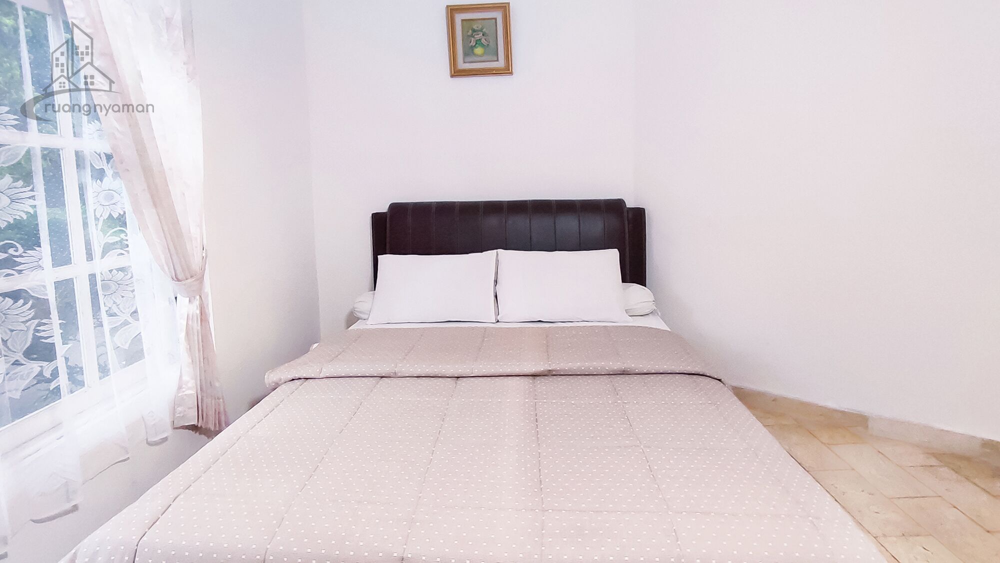 Cama bem arrumada com colcha de poá e dois travesseiros, janela com cortinas florais e um quadro na parede.