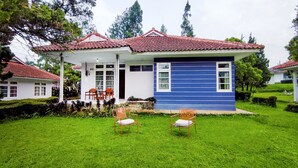 A two-story house with a red-tiled roof, a blue exterior wall, and a front porch with two wooden chairs. - Ruang Nyaman at Sukanagalih Resort (Cipanas)