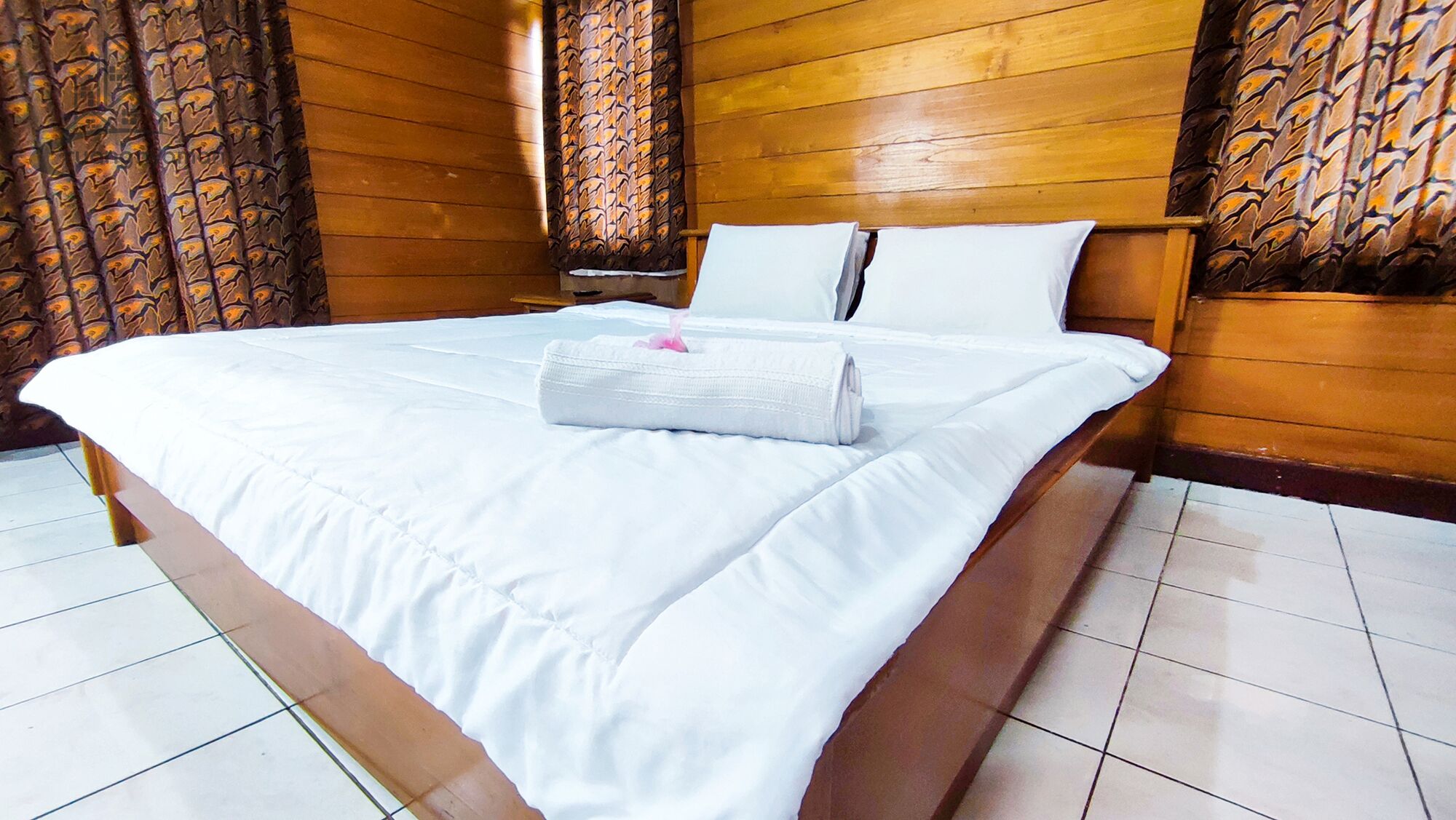 A neatly made bed with white linens and a rolled towel, wooden headboard, and patterned curtains.