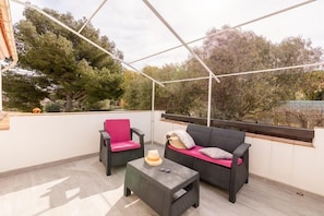 A covered outdoor seating area with two wicker sofas and a small table, surrounded by a white wall and a view of trees.