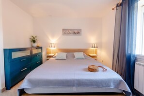 Ein Schlafzimmer mit Bett, Nachttischen, einem Schminktisch, einer Wandlampe und einem Fenster mit Vorhängen.
