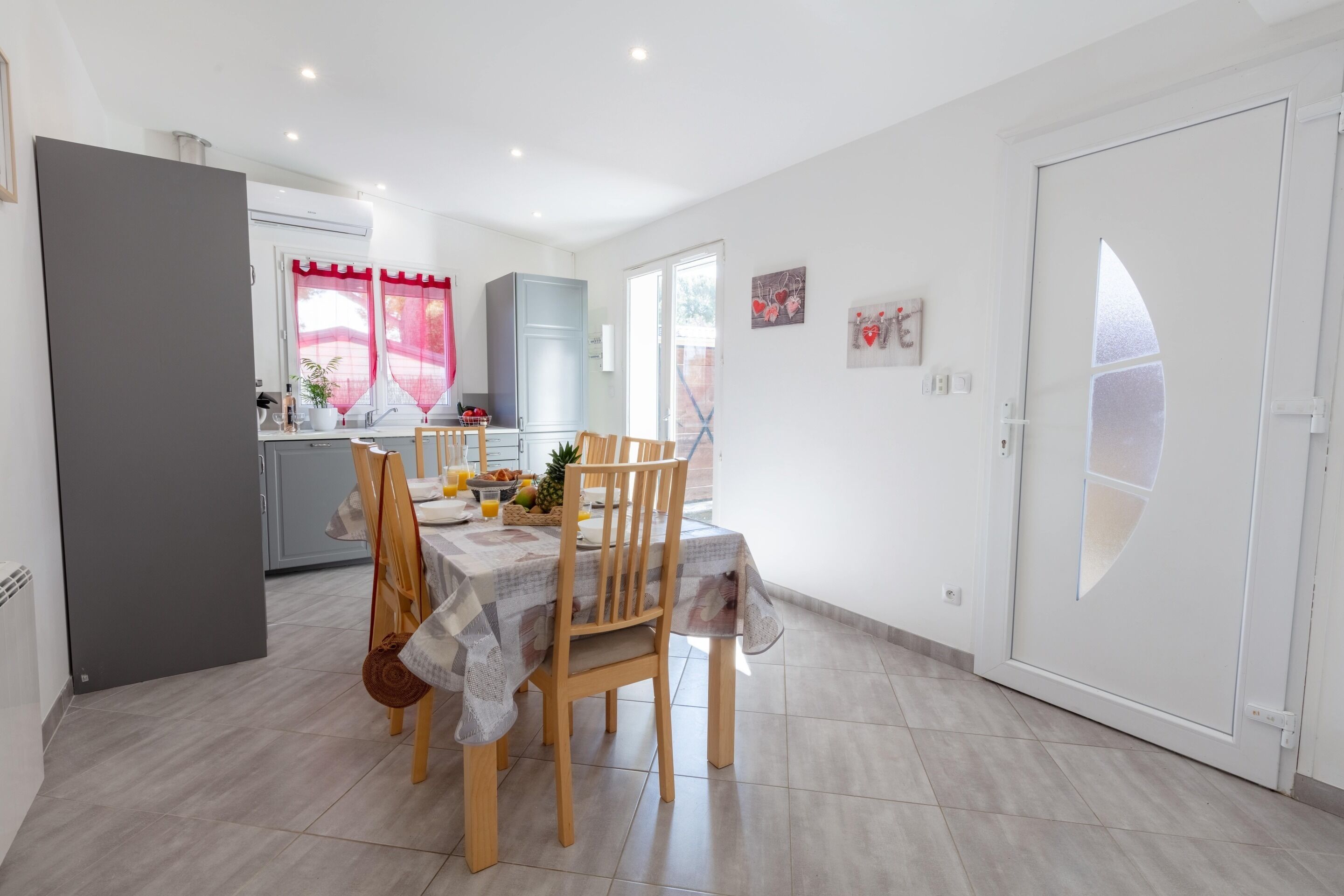 A modern kitchen with a dining area, featuring a table set for two with chairs, a refrigerator, and a window with red curtains.