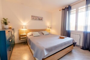 Een slaapkamer met een bed, nachtlampen, een nachtkastje, een dressoir, een raam met gordijnen en een aan de muur hangend schilderij.
