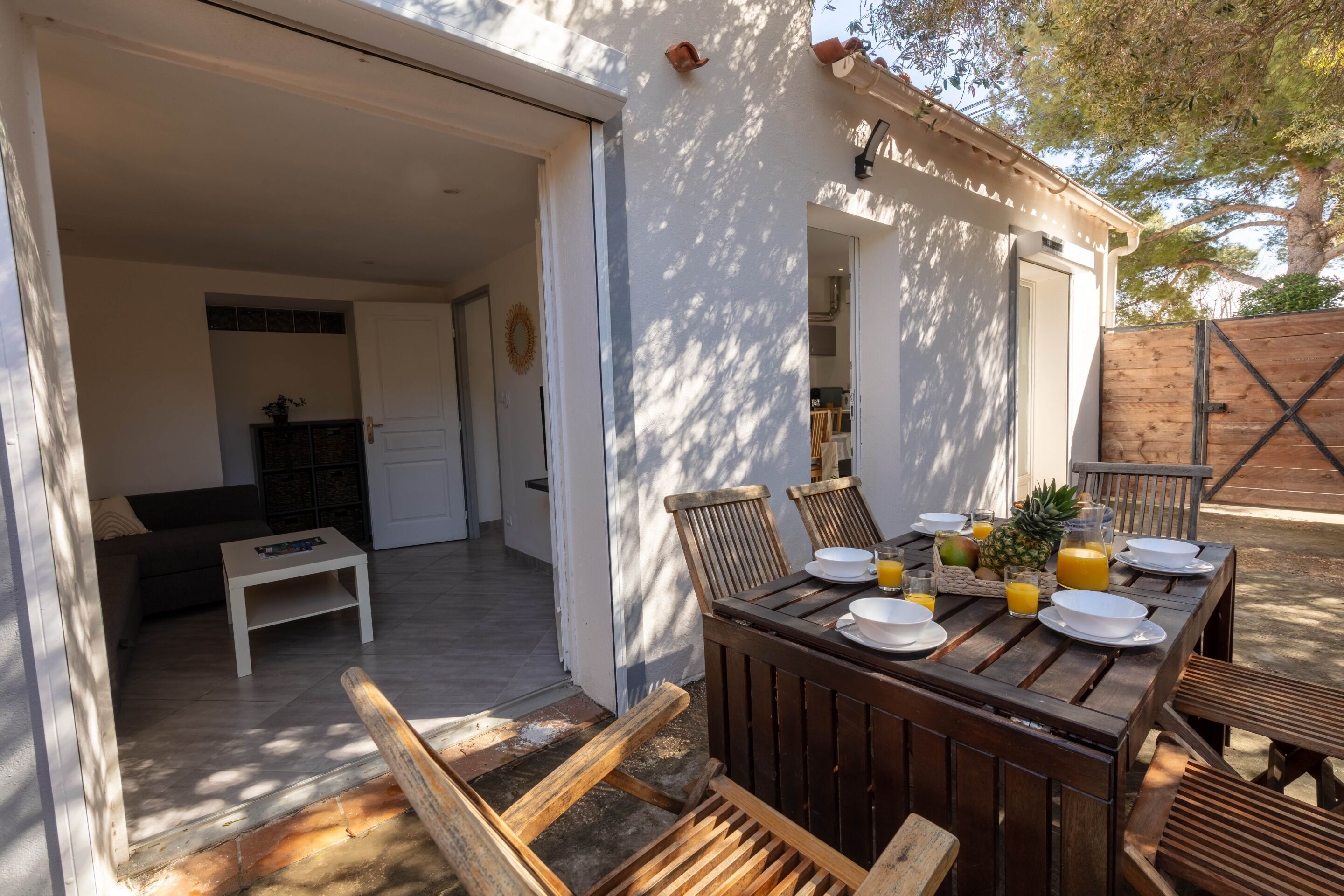 A wooden dining table set with plates, glasses, and a pineapple, with a view of a kitchen and a patio area.