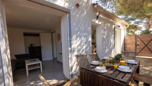 Une table de salle à manger en bois, dressée avec des assiettes, des verres et un ananas, avec vue sur une cuisine et un espace patio.