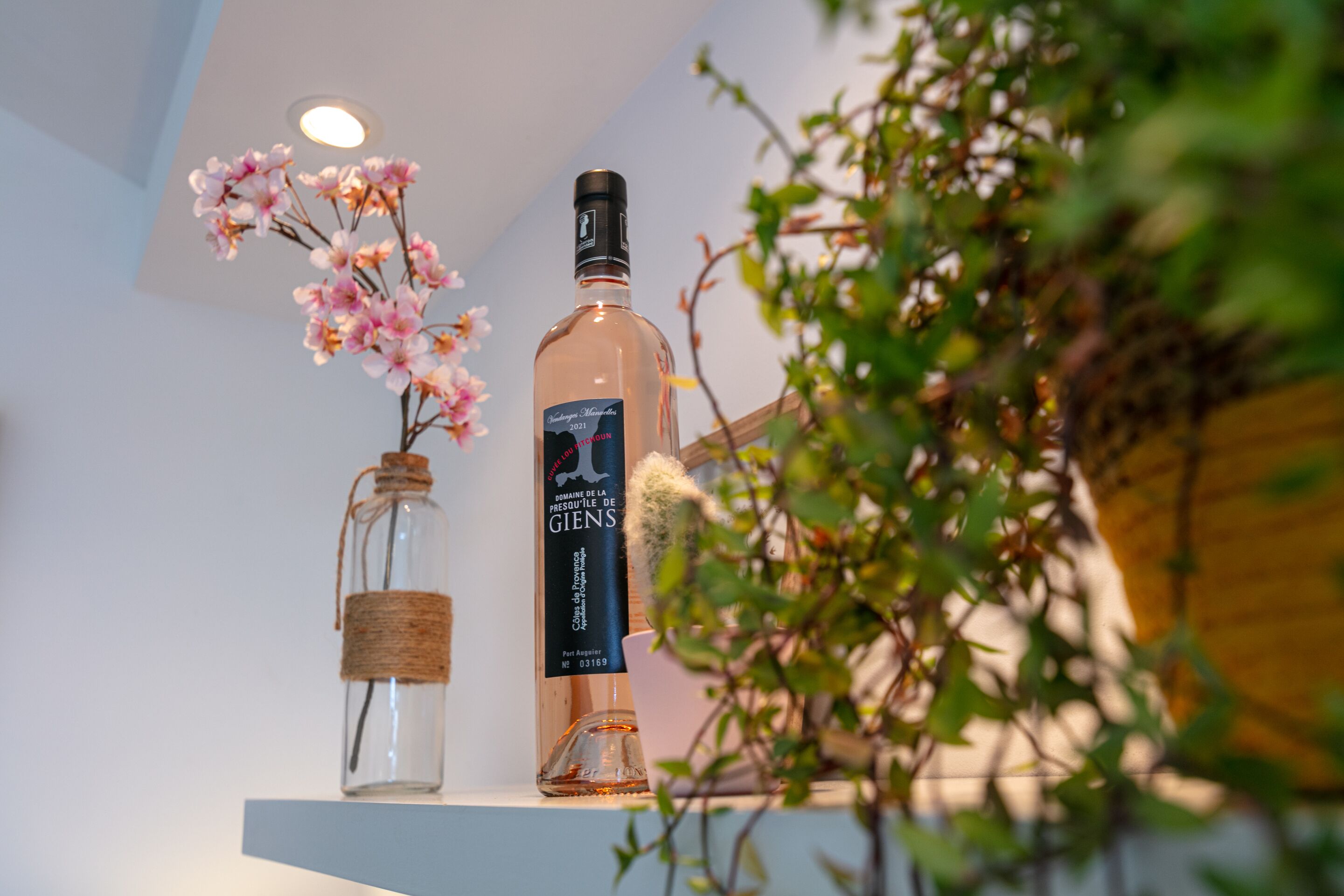 Una botella de vino con etiqueta, un recipiente de vidrio con corcho y flores rosadas sobre una repisa.