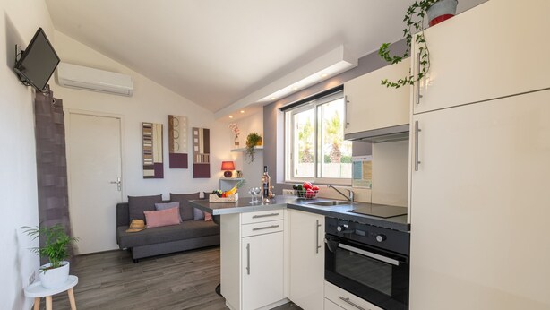 A compact kitchen with a built-in oven, a dining area with a sofa, and a window with a view of palm trees.