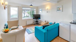 Uma sala de estar moderna com um sofá azul, uma mesa de jantar com frutas, uma televisão e uma pequena cozinha.