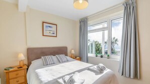 A neatly made bed with pillows and a folded towel, a bedside table with a lamp, a window with curtains, and a framed picture on the wall. - Amazing Sea Views from this Ground Floor 2 Bedroom High Spec Apartment (Torpoint)
