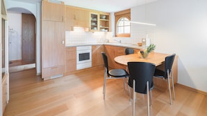 A modern kitchen with wooden cabinets, a built-in oven, and a dining area with a round table and four chairs. - Haus Bärgbach by Zermatt Premium Apartments (Zermatt)