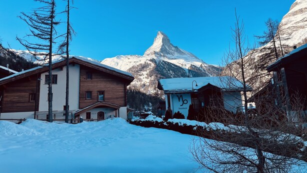 A wooden cabin with a snow-covered roof, a small adjacent building with graffiti, and a mountain with a distinctive peak in the background. - Haus Bärgbach by Zermatt Premium Apartments (Zermatt)