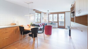 A modern kitchen with wooden cabinets, a dining area with a table and chairs, and a red accent chair. - Haus Bärgbach by Zermatt Premium Apartments (Zermatt)