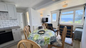 A compact kitchen with white cabinets, a dining table with a leaf-patterned tablecloth, wooden chairs, and a view of the sea through large windows.