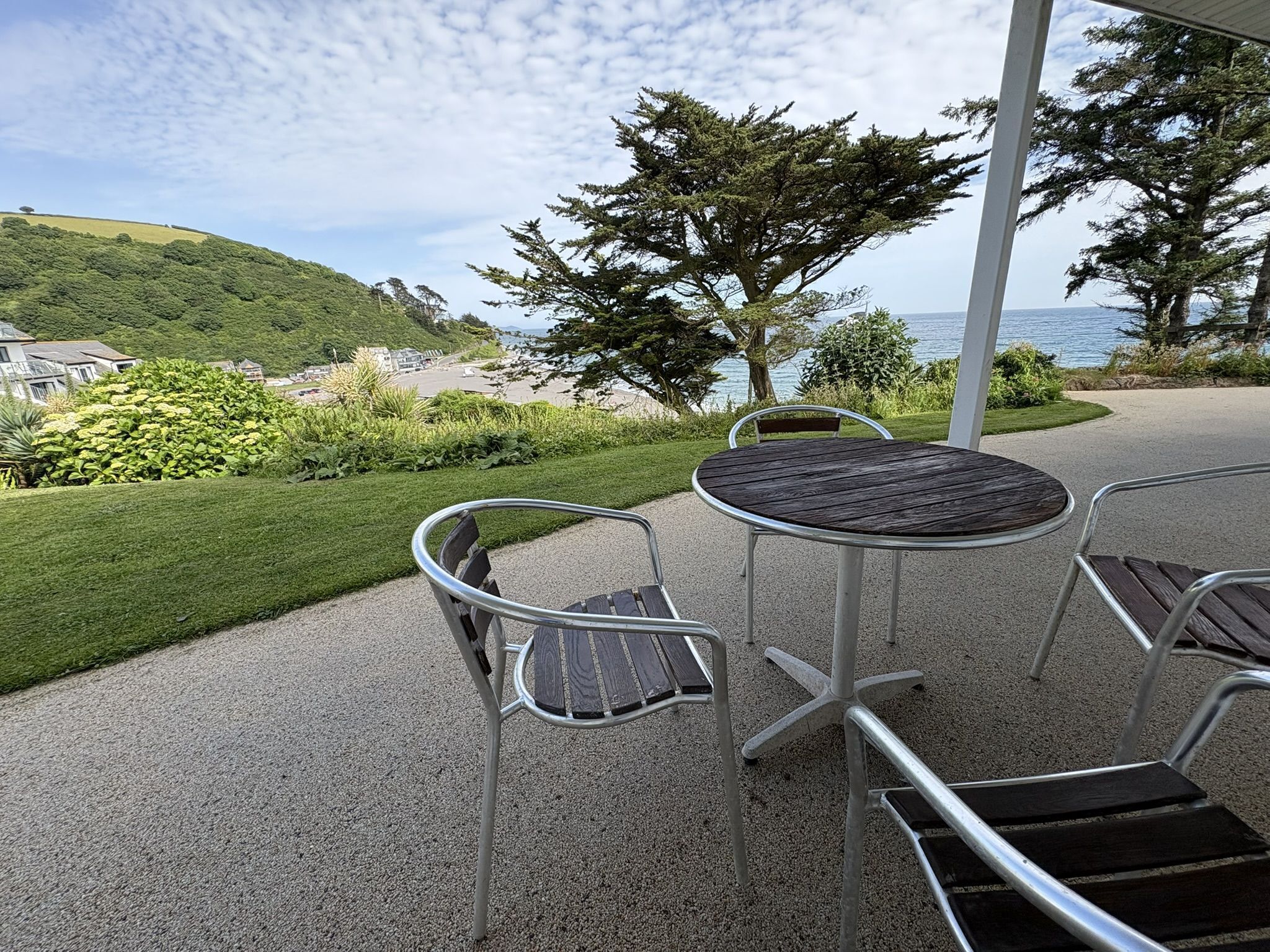 Um pátio externo com uma mesa redonda e quatro cadeiras, com vista para uma paisagem costeira com colinas e o mar.