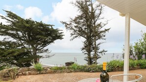 Ein Tisch mit einer Weinflasche und Gläsern, umgeben von Stühlen, mit Blick auf das Meer und Bäume.