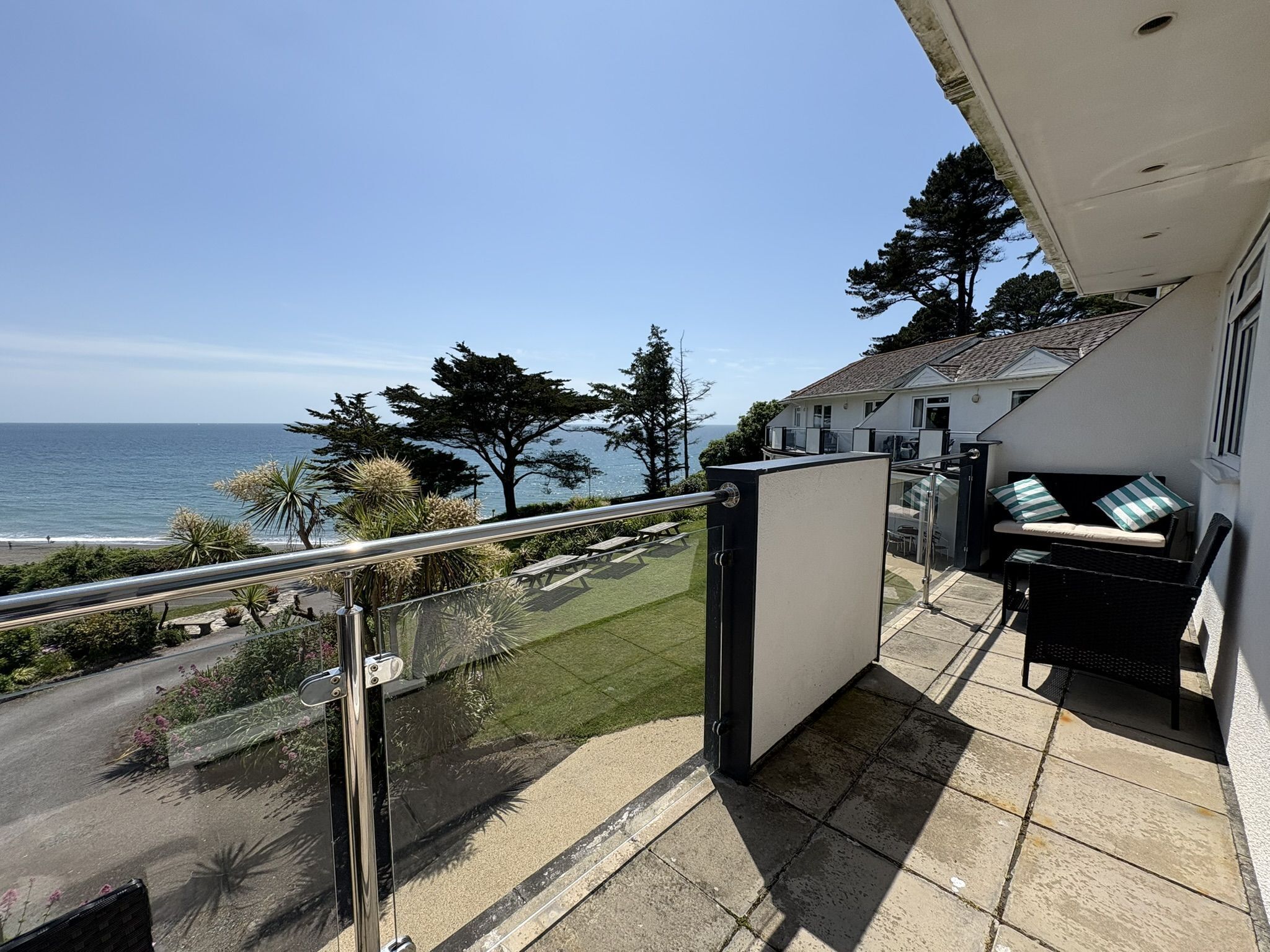 A balcony with a view of the ocean, a grassy area, and a building with a white exterior.