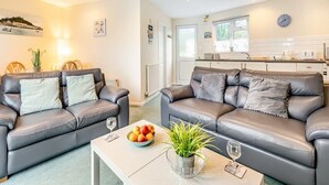 A modern living room with two leather sofas, a coffee table with fruit and a plant, and a kitchen in the background. - Cornish Apartment with Private Balcony & Stunning Sea Views (Seaton)