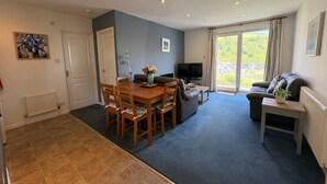 Een woonkamer met een eettafel, stoelen, een bank, een televisie en uitzicht op een groen landschap door grote ramen.