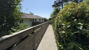 A pathway leading to a white building with a sloped roof, surrounded by dense greenery and a wooden fence. - Cosy Apartment with Private Balcony & Fantastic Sea Views (Torpoint)