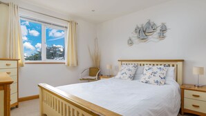 A bedroom with a wooden bed, a nightstand, a chair, a window with curtains, and a wall decoration. - Cornish Duplex 2 Bedroom Apartment with Stunning Sea Views (Torpoint)